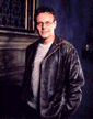 Tony in brown leather jacket, standing in front of fireplace